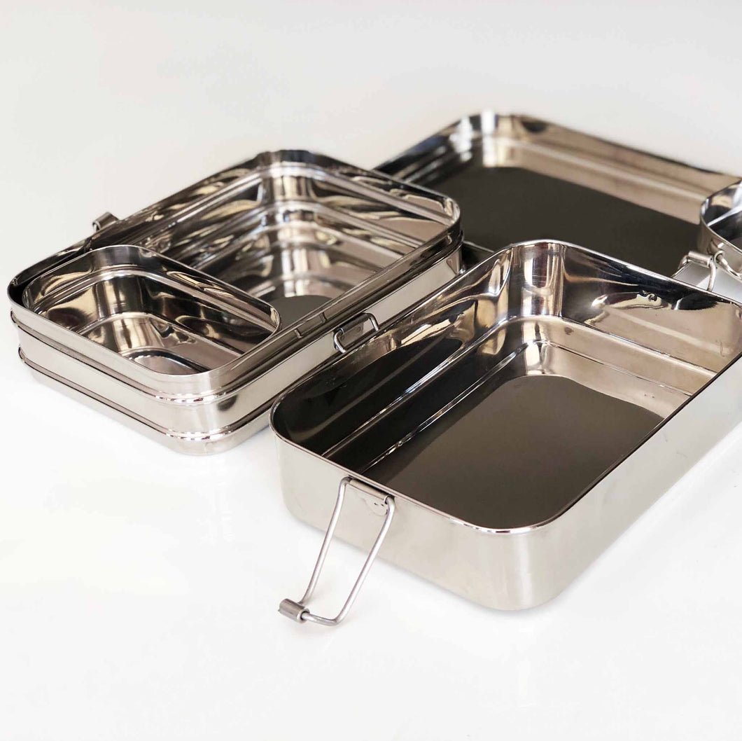 Stylish double layered stainless-steel lunchbox by Bento Ninja, seen here with all the pieces laying side by side with clasps open and the snack box open and sitting inside the top layer of the lunchbox.