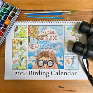 This 2024 Birding Calendar is the unofficial field guide to New Zealand birds. Seen here on a wooden table surrounded by paints, paintbrushes and binoculars.