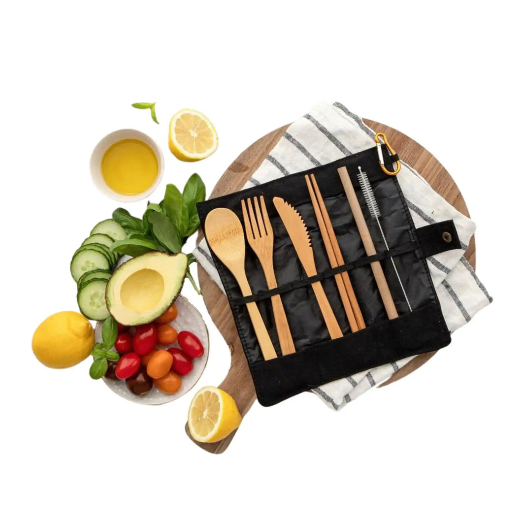 Bamboo cutlery roll with knife, fork, spoon, chopsticks, straw and cleaning brush in the unrolled or open position sitting on a tea towel on top of a wooden chopping board with avocado, lemon, tomato, cucumber, basil and other salad ingredients nearby on a white background.