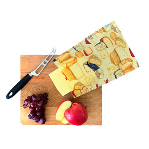 Beeswax cheese bag with cheese block pattern on a wooden chopping board with grapes. sliced apple and a cheese knife.