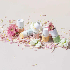 Eco friendly lip balms in compostable cardboard tubes seen here in a line with some on an angle and some open on their sides surrounded by petals and flowers with a very light pink background.