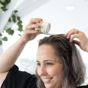Dry shampoo for light hair, scented with Lime and sweet orange essential oils. Seen here in use, a woman with shoulder length hair is shaking the tube into her roots and fluffing with her other hand. White background with some green leaves.