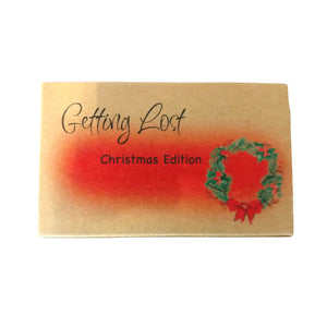 The Christmas edition of the Getting Lost Adventure game in a kraft cardboard box with an orange cloud and a Christmas wreath with a red ribbon on the front.