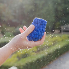 A blue EcoSplat reusable water balloon which looks like it has just been caught in a hand with droplets of water flying everywhere and the blurry greenery of a garden in the background.