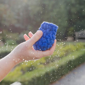 A blue EcoSplat reusable water balloon which looks like it has just been caught in a hand with droplets of water flying everywhere and the blurry greenery of a garden in the background.