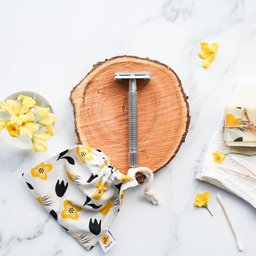 Stainless steel safety razor displayed on a cross section of a tree, flowers and a Kowhai bag sit around the razor.