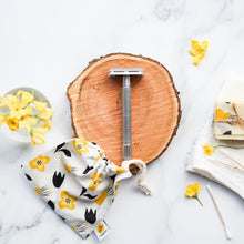 Stainless steel safety razor on the cross section of a tree with a draw-string tote back in the foreground and flowers dotted about.