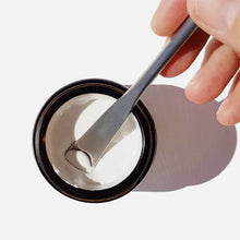 Fingers in the top right hand corner of the image are holding a stainless steel spatula which is scooping toothpaste from a glass jar, white background and shadow cast to the right of the jar.