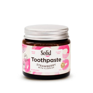 Solid Oral Care strawberry toothpaste with fluoride in an amber glass jar and aluminium lid. The white label with pink swirls reads made in NZ and plastic free.