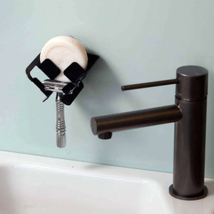 The Block Dock vertical soap dish and safety razor holder seen here attached to a wall above a sink next to a black tap. The Razor Dock is black with a circular white soap and a silver safety razor in front.